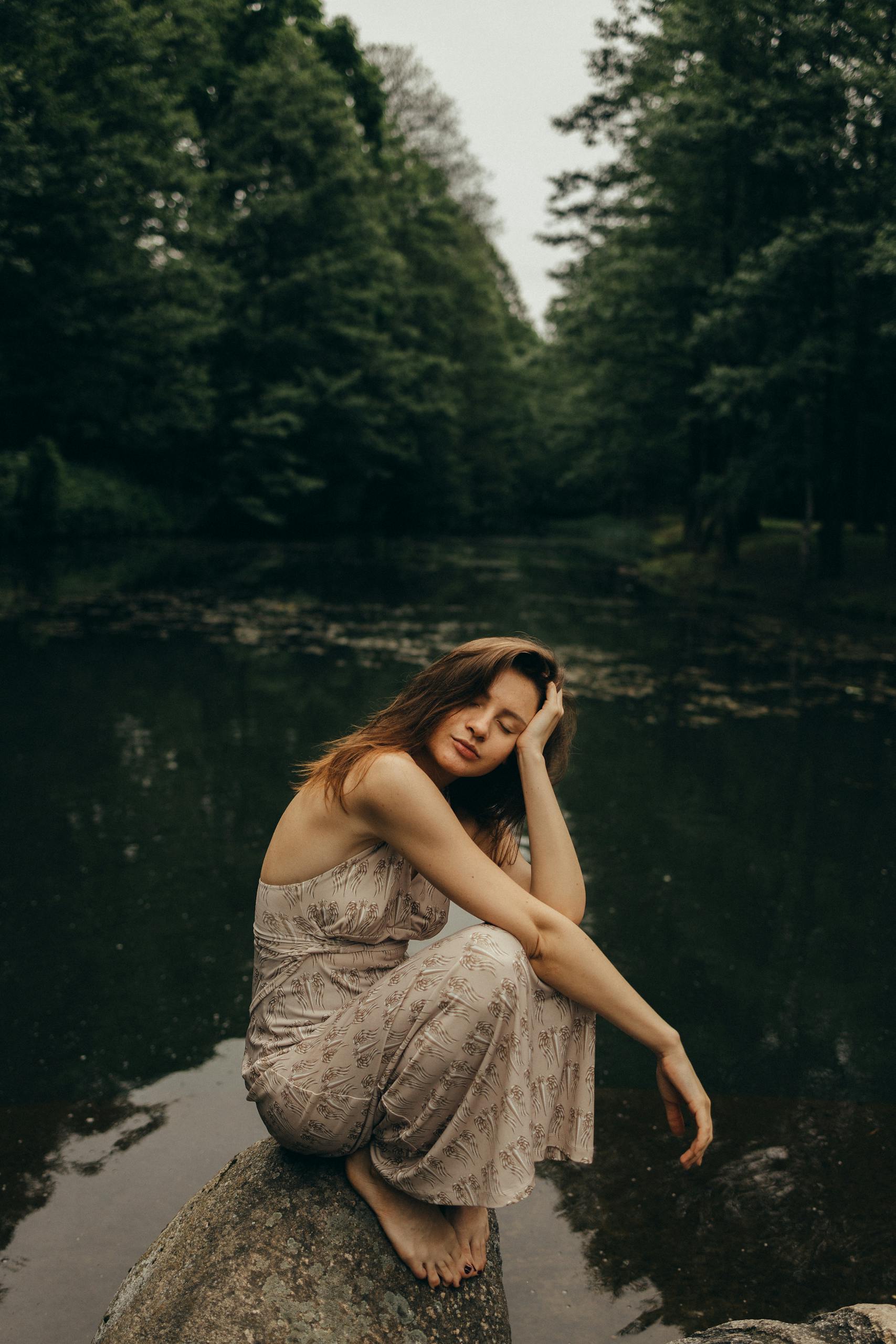 A woman in a dress sits peacefully by a calm river in the forest, capturing a moment of solitude and nature.